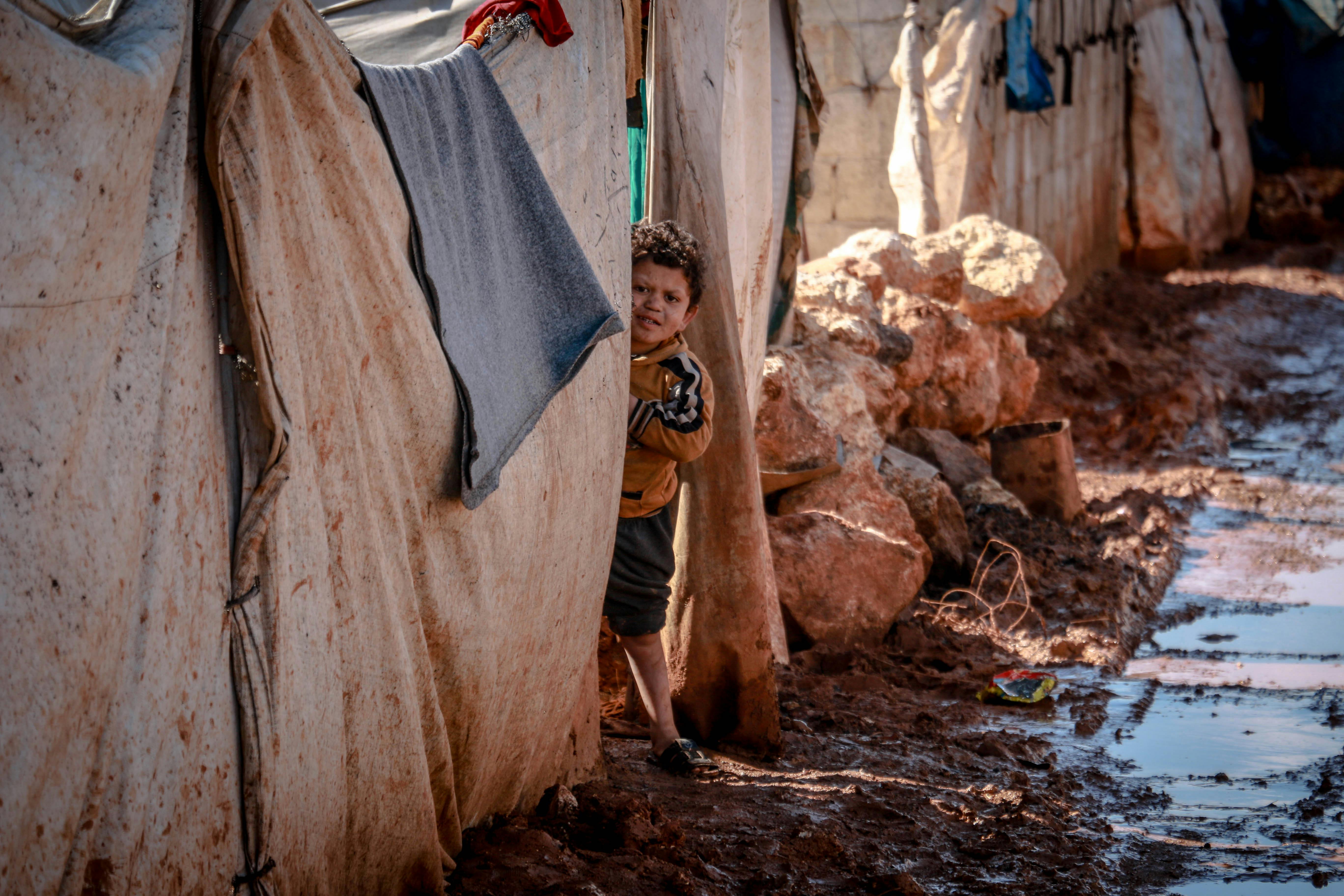 Child standing beside a temporary shelter in a muddy refugee camp during Middle East conflict, highlighting humanitarian impact amid Iran–Israel tensions and regional instability.